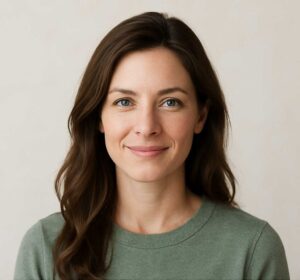 A portrait of Elena Hart, a wellness coach and yoga practitioner with chestnut-brown hair and blue eyes, smiling warmly in a sage green sweater against a soft, neutral background.